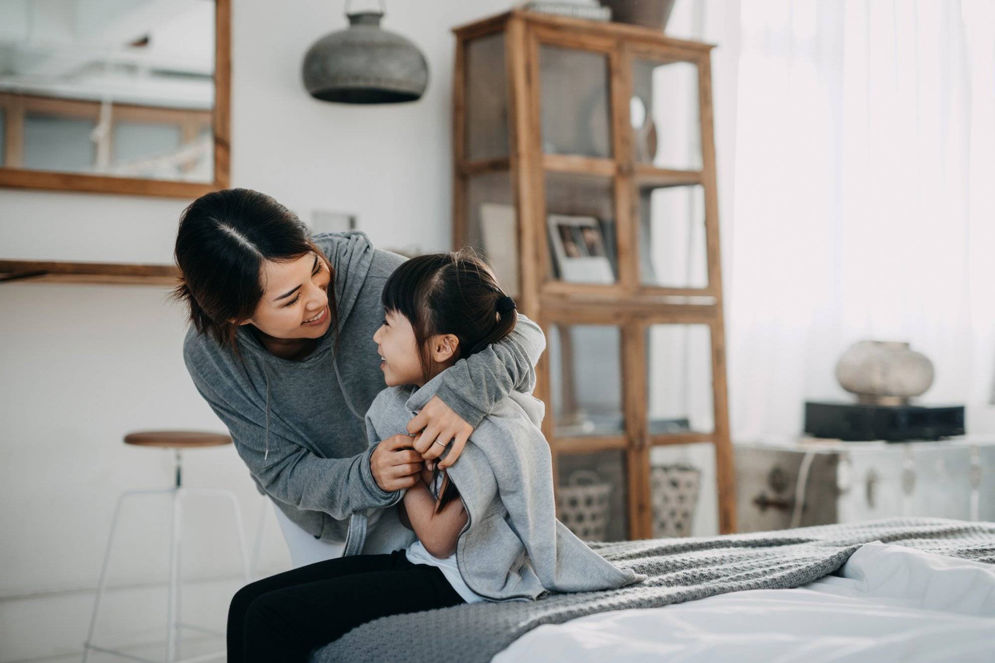 Caring young Asian mother putting a coat on her daughter at home